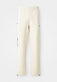 Light beige sweatpants made from soft fabric. Features a black logo, side pockets with zippers, and a relaxed fit.