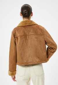 Veste en suède synthétique marron avec un large col et un bord en fourrure, dotée de longues manches et d'une coupe courte, associée à un jean de couleur claire.