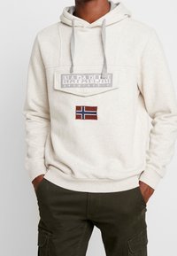 Sudadera color beige con un bolsillo frontal, cordones grises y una bandera de Noruega bordada. El material parece suave y cómodo.