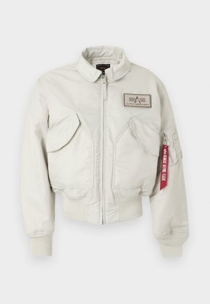 Hellbeige Bomberjacke mit Frontreißverschluss, zwei großen schrägen Taschen, Alpha Industries-Logo auf der Brust und rotem "Remove Before Flight"-Anhänger am Reißverschluss des Ärmels.