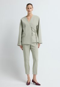 Jasnozielona kurtka z wiązaniem, długimi rękawami i kieszenią z przodu, w połączeniu z dopasowanymi spodniami i bordowymi szpilkami.