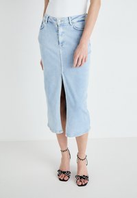 Light blue denim midi skirt with a front slit, fitted design, and belt loops. Paired with black sandals featuring white polka dots and bows.