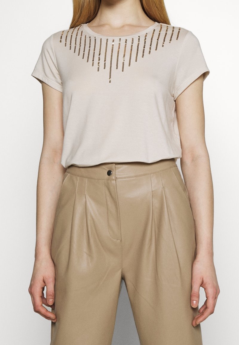 Haut beige à manches courtes avec des rayures verticales en sequins dorés, associé à un pantalon en cuir plissé de couleur camel, taille haute.