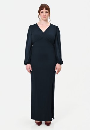 Donna con capelli rossi che indossa un lungo vestito nero con maniche lunghe e scarpe bordeaux con punta, in piedi su uno sfondo chiaro e uniforme.