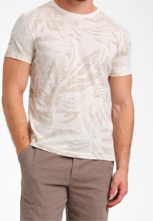 Mężczyzna ubrany w beżowy T-shirt z krótkim rękawem w wzory oraz brązowe spodnie z fakturą, z jedną ręką w kieszeni na białym jednolitym tle.