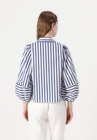 Valkoinen ja siniraitainen pusero, jossa on puffihihoja, kauluspäällinen ja nappi yksityiskohtana takana. Kangas on sileä.