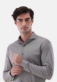 Camicia grigia a maniche lunghe con un sottile motivo a trama, chiusura frontale con bottoni e colletto classico. Presenta bottoni scuri e polsini aderenti.