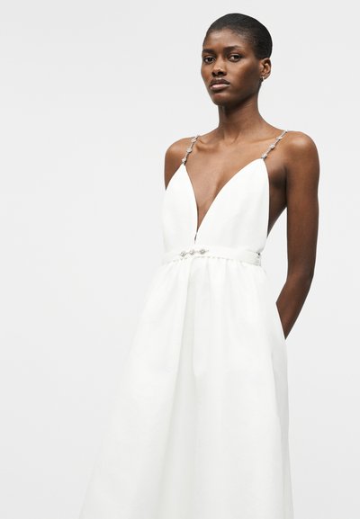 Γυναίκα που φοράει ένα λευκό φόρεμα χωρίς μανίκια με βαθύ V λαιμόκοψη, διακοσμητικές τιράντες και διακοσμητική ζώνη στη μέση.