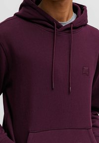 Marinröd hoodie i mjukt tyg, med en dragsnodd på huvan, känguruficka och en liten logotyp på bröstet.