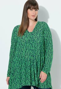 Blusa floreale verde con scollo a V, maniche lunghe e vestibilità ampia. Presenta motivi floreali blu scuro e una texture del tessuto morbida.