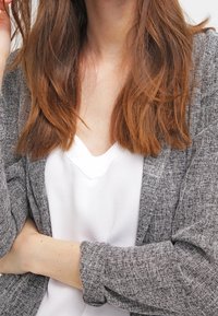 Cardigan gris texturé porté sur un haut blanc à col en V, mettant en valeur un look décontracté en couches. Le cardigan possède des manches longues et une coupe décontractée.