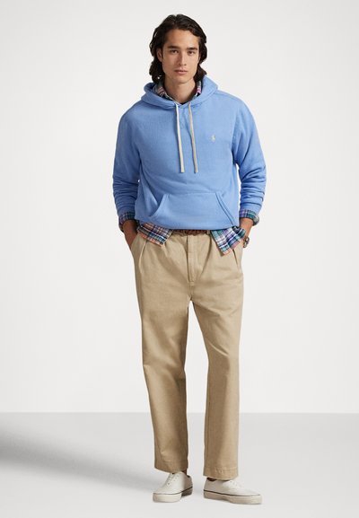 Ανοιχτό μπλε φούτερ με κορδόνια, τσέπη μπροστά, φορεμένο πάνω από πουκάμισο καρό. Μπεζ chinos και λευκά αθλητικά παπούτσια ολοκληρώνουν την εμφάνιση.