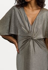 Třpytivé stříbrné šaty s výstřihem do V, uzlovým detailem vpředu a širokými, volnými rukávy. Jsou vyrobené z texturované metalické tkaniny.