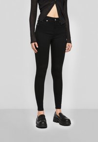 Calças skinny pretas em material elástico, com uma cintura alta, design de cinco bolsos e acessórios em metal, combinadas com mocassins pretos chunky.