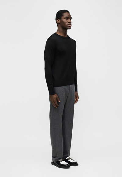 Homme debout de profil portant une chemise noire à manches longues, un pantalon gris et des mocassins en cuir noir brillant avec des détails métalliques.