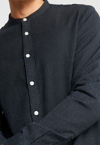 Camicia button-up in tessuto blu navy con pois bianchi. Presenta un colletto alzato e bottoni simili a perle. Dettaglio in tessuto texturizzato.