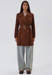 Trench coat marrone con cintura, ampi rever e due tasche frontali, abbinato a pantaloni in denim chiaro e larghi e calzature marroni.