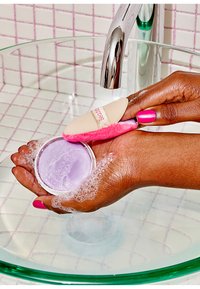 Dłoń trzyma lawendowy balsam oczyszczający w okrągłym pojemniku z bąbelkami. Różowo-beżowa gąbka aplikacyjna jest używana do czyszczenia nad szklaną miską.