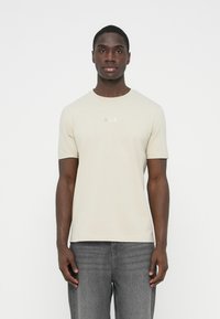 Svetlo bež kratka majica s kratkimi rokavi, okroglim izrezom in subtilnim logotipom "BOSS" v kovinskem sijaju. Kombinirana s temno sivimi kavbojkami.