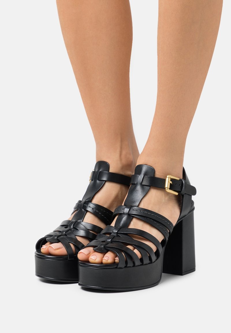 Black leather platform sandals with a woven design, wide straps, and a bold block heel. Features a metal buckle on the ankle strap.