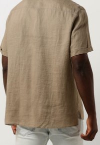 Persona di pelle scura che indossa una camicia in lino beige a maniche corte e jeans azzurri, vista di spalle su uno sfondo semplice.
