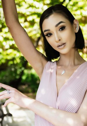 Model wearing a pleated light pink top, silver leaf necklace, and rings, with short dark hair and raised arm against a green background.