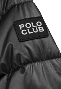 Czarna tkanina kurtki puchowej o gładkiej, błyszczącej fakturze; posiada prostokątną czarną metkę z białym wypukłym napisem "POLO CLUB".