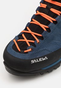 Niebieskie zamszowe buty trekkingowe z czarnym materiałem tekstylnym i jaskrawymi pomarańczowymi sznurowadłami. Posiadają solidną czarną gumową podeszwę z teksturowanym gripem. Logo po boku.