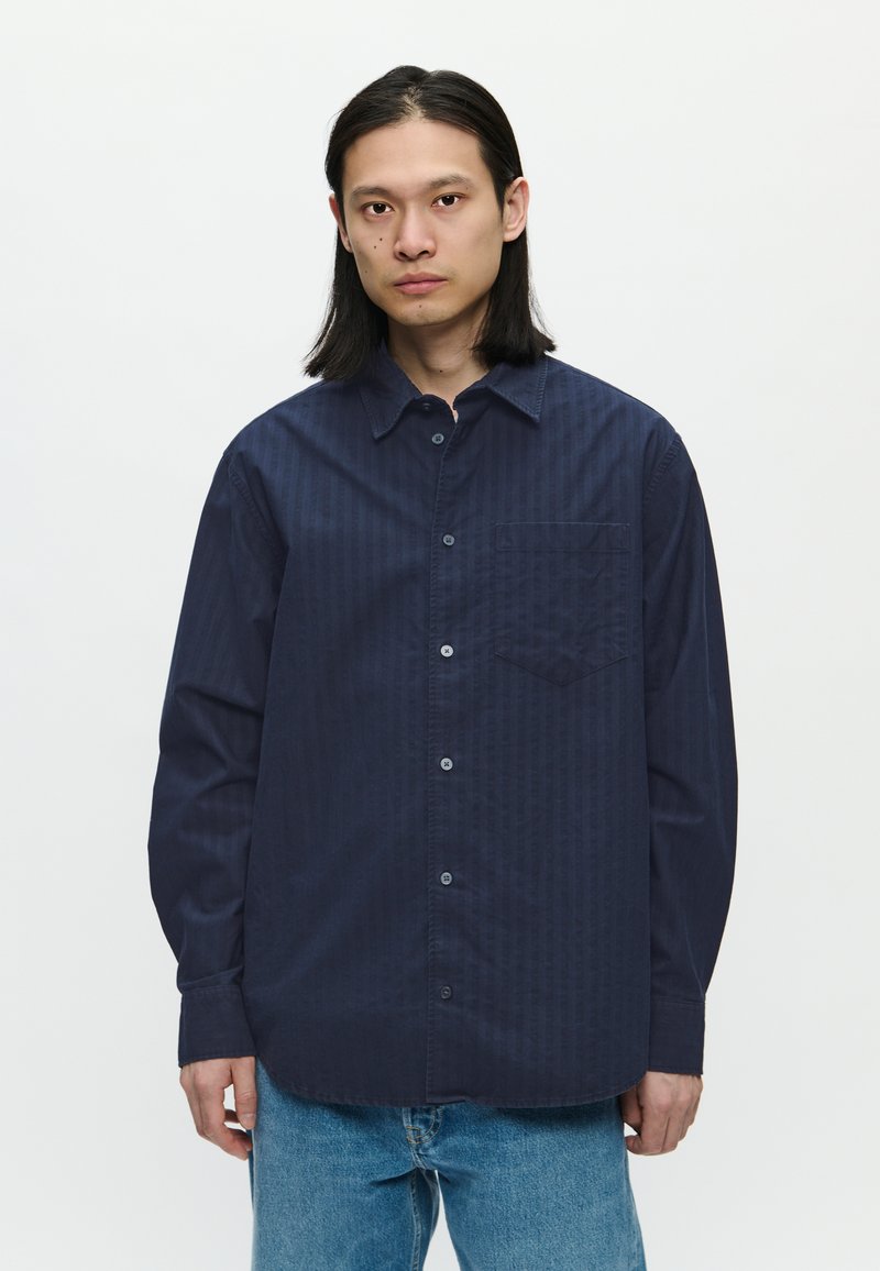 Camisa de botón azul oscuro, con rayas verticales, de mangas largas y un bolsillo en el pecho. La tela parece suave con una textura sutil.