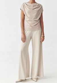 Vaaleanbeige drapattu yläosa lyhyillä hihoilla, yhdistetty leveälahkeisiin vaaleanbeigeihin housuihin, valmistettu sileästä kankaasta; mallilla on päällään terävät vaaleanbeigeit kengät.