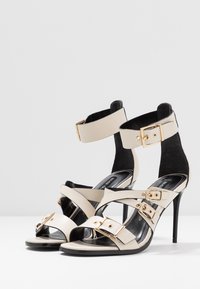 Sandalias de tacón alto beige con correas de cuero entrelazadas, hebillas de metal dorado y una correa de tobillo de ante negro. Acabado suave, diseño de punta afilada.