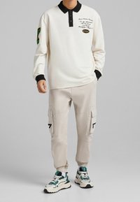 Muž v off-white polo tričku s dlouhým rukávem, černým límcem a manžetami, béžových cargo kalhotách a bílých teniskách s zelenými a černými akcenty.