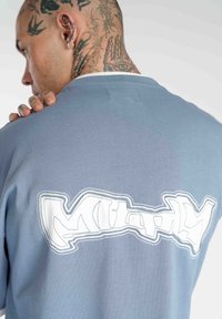 Blå bomulds T-shirt med et stort hvidt graffiti-inspireret logo på ryggen. Stoffet ser blødt ud og har en afslappet pasform.