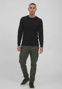 Schwarzer gerippter Strickpullover kombiniert mit olivgrünen Cargo-Hosen und schwarzen Sneakern. Die Cargo-Hosen verfügen über mehrere Taschen.