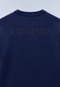 Πίσω μέρος μπλε ναυτικού πουκαμίσου με παραμορφωμένο μαύρο κείμενο που εκτείνεται οριζόντια κοντά στον λαιμό.