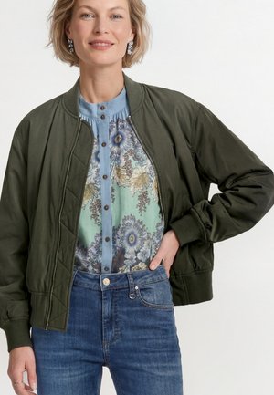 Frau trägt eine olivgrüne Bomberjacke über einem blauen, geblümten Hemd mit Knöpfen und blauen Jeans, posiert mit einer Hand auf der Hüfte.