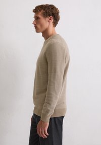 Pull beige en maille avec col rond et poignets côtelés, tissu texturé, silhouette ajustée, associé à un pantalon gris foncé.