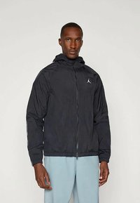 Schwarze leichte Jacke mit Kapuze, Reißverschluss vorne, elastischen Bündchen und Saum. Mit einem weißen Logo auf der linken Brust. Glatte Textur.