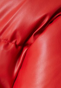 Detail červené kůže, která ukazuje hladký a texturovaný povrch s jemnými vráskami a švy, zdůrazňující kvalitu a provedení materiálu.