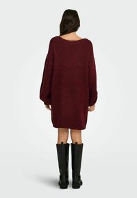 Przewymiarowana bordowa dzianinowa sukienka swetrowa z szerokim dekoltem i długimi rękawami, prezentowana z czarnymi kozakami do kolan. Teksturowana tkanina.