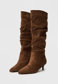 Par de botas altas de ante marrón, con diseño suelto, puntas afiladas y tacones kitten, sobre fondo blanco.