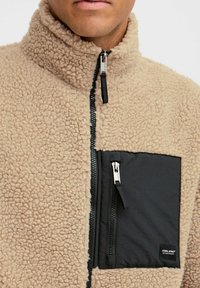 Beige fleece jacket with a high collar, black zippered chest pocket, textured material, and contrasting black zipper detailing.