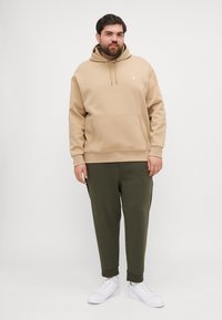 Beige hoodie met kangoeroezak, trekkoordcapuchon en klein logo, gecombineerd met donkergroene sweatpants en witte sneakers.