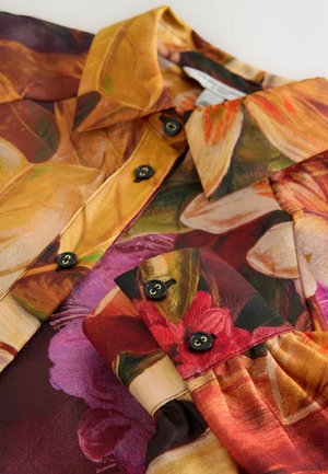 Bluse mit floralem Muster in warmen Tönen von Gelb, Rot und Bordeaux; mit schwarzen Knöpfen und einem strukturierten Kragen; glatte Stofftextur.