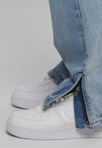 Zapatillas blancas de cuero con perforaciones, combinadas con unos jeans de denim azul claro que presentan un zipper lateral y bajos ligeramente desgastados.