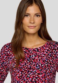 Frau mit langen braunen Haaren, die ein farbenfrohes Oberteil mit rot, lila, schwarz und weißem Muster vor einem hellgrauen Hintergrund trägt.