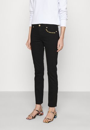 Persona con jeans negros ajustados de corte slim con botón dorado y texto "Versace Jeans Couture", camiseta blanca de manga larga y sandalias coloridas con tiras.