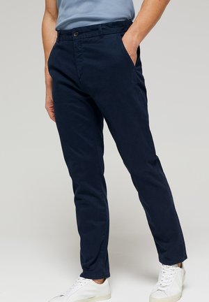 Navy blue chino pants with a tailored fit, made of cotton fabric, featuring front pockets and a button closure. Matched with white sneakers.