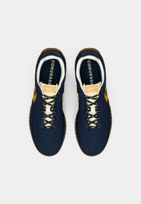 Marineblå ruskind sneakers med sorte og hvide snørebånd, udstyret med en gummisål og et gult Converse-logo på tungen.