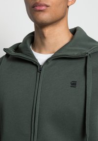 Sweat à capuche zippé ajusté vert avec un col haut, une fermeture éclair contrastante et un petit logo noir sur la poitrine. Tissu doux et texturé.
